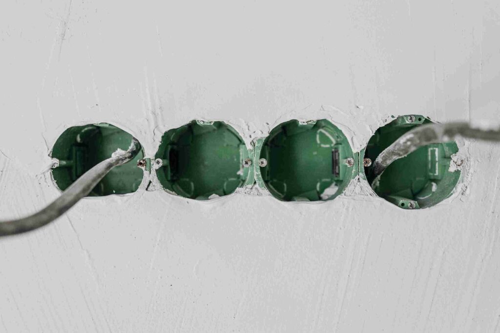 Close-up of green electrical outlet boxes in a damaged white wall with torn plaster and a cable running through them.