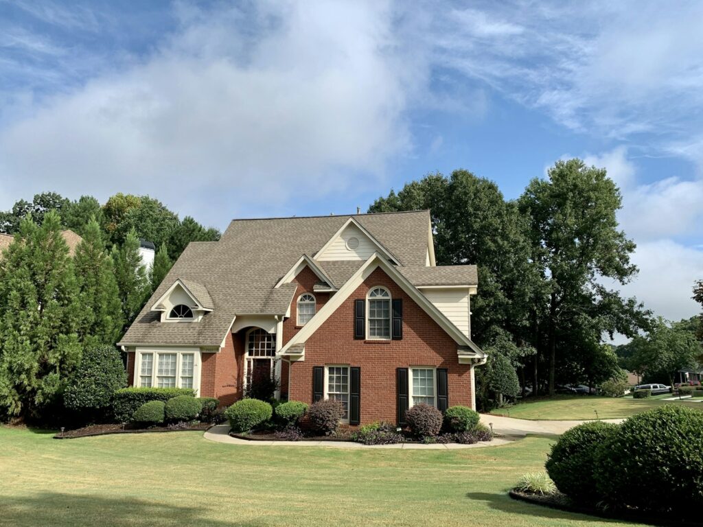 Front view of a brick suburban house with gabled roof, arched windows, and neatly trimmed shrubs in the yard under a blue sky.