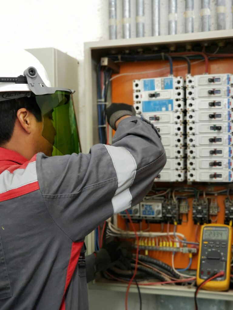 Electrician in a hard hat and face shield testing an electrical panel