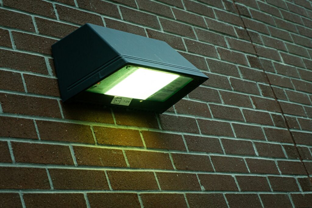 Exterior wall-mounted black floodlight on brick wall emitting bright white light onto the wall below