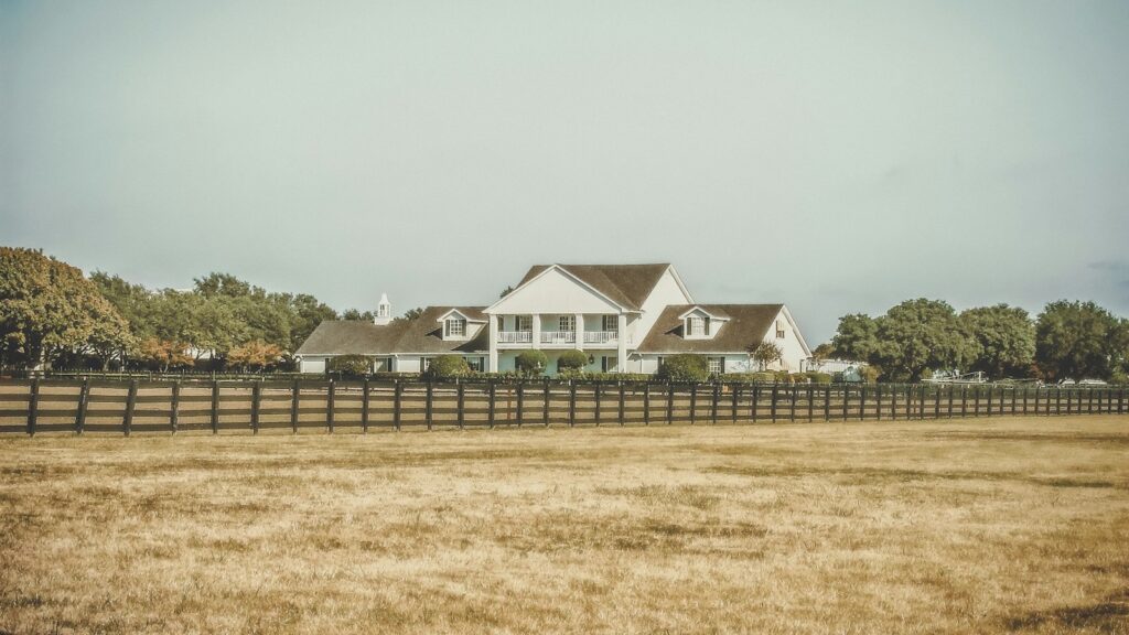 White farmhouse with a steep roof behind a dark wooden fence in a dry, open field under a pale blue sky.